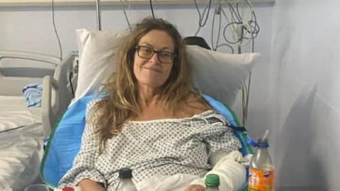 A woman with long blonde hair and glasses is is lying in a hospital bed, wearing a hospital gown. There are drinks in front of her and wires behind connected to the bed.