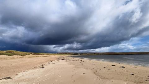 A sandy beach stretches into the distance with grassy dunes to the left and the sea to the right. Above a huge dark grey and black cloud stretches across the sky.