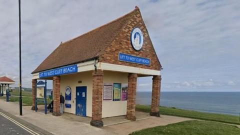 Whitby Cliff Lift - a red-brick building with a pointed roof and blue signage reading 'Lift to West Cliff Beach'. The building is overlooking a view of the sea.