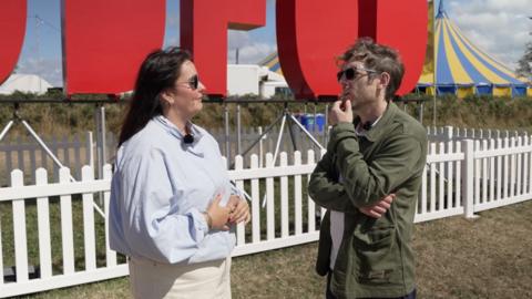 In this picture there are a man and woman in glasses looking at one another. The man has a green jacket with a white t-shirt on, the woman is wearing a light blue jacket and cream trousers. There is a white fence in the background, along with a blue and yellow tent and large letters that are mostly cut off from the frame spelling Eisteddfod. 