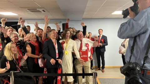 Jessica Toale wins Bournemouth West - she is wearing a white, fitted, long-sleeved dress and a red Labour rosette, and appears with other Labour Party members, all smiling broadly and at camera to the right of the frame