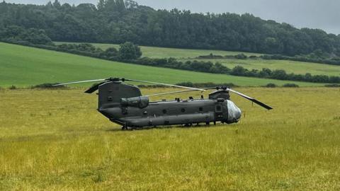 A chinook landed in a field, it's a dark grey colour with two large propellers surrounded by green grass field