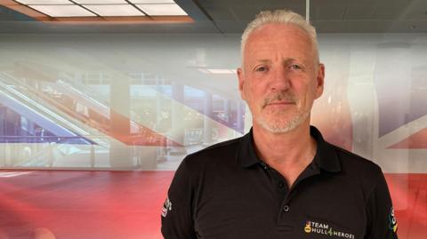 Head and shoulders shot of a man with grey hair and goatee beard standing in front of a Union flag. He is wearing a black polo shirt and has his arms behind his back.