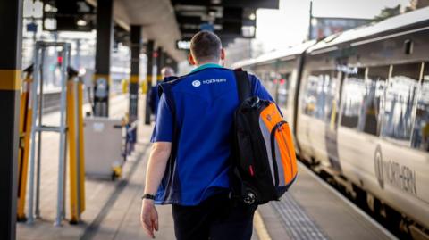 A Northern Rail conductor walks down the platform with his back facing the camera. He is wearing a blue Northern Rail jacket, with a Northern Rail train to his right