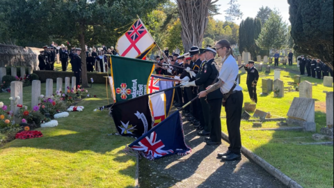 A row of people in uniforms in a cemetery at low light, surrounded by trees. They stand on a path holding flags at different angles as they raise them. In front of them a line of grave stones with flowers and wreaths in front of them. Other people in military and church uniforms stand behind them.