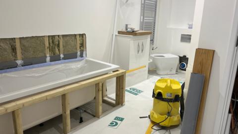 bathroom that is half complete, the bath is on the left with a tap and basin in the background. there is a yellow vacuum cleaner on the right with wood and piping.