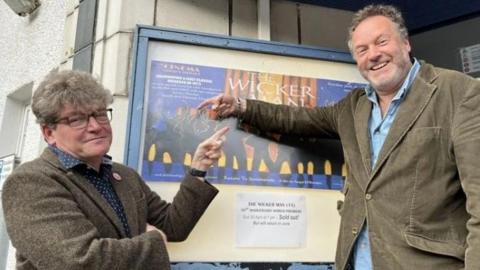 Two men in their 50s either side of a poster for The Wicker Man outside a cinema. Dominic, on the left, is in a tweed suit and has glasses and thick hair. Justin (right) is in a cord suit and has a beard. They are smiling and pointing at the poster.