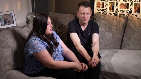 A woman in a blue t-shirt and a man in a black t-shirt sit on a sofa