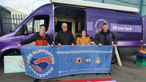 Four people stand holding up a blue banner which reads 'Fans supporting foodbanks' and 'hunger doesn't wear club colours'. They are standing in front of a purple van. 