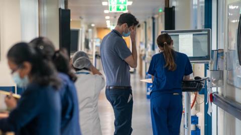 Generic view of two medical staff members from behind looking at a laptop. Some other people in scrubs are seen blurred walking around them.