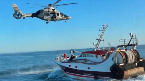 A helicopter is hovering above a white trawler. A line can just be seen at the front of the boat beside a person wearing a harness and red clothing.