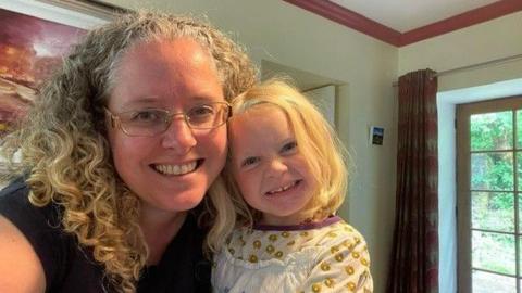 A woman with glasses and curly long blonde hair cuddles into her daughter who has blonde straighter hair and is grinning widely