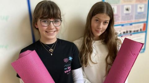 Two girls stand against a wall smiling and holding pink yoga mats