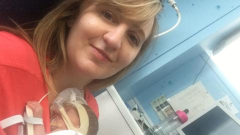 A woman with blonde hair in hospital cradling a very young baby