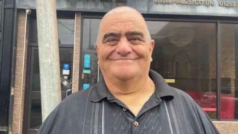 A picture of a smiling Brian Roberts. He is a larger man with a balding head and is wearing a black striped polo shirt. He is standing in front of a former restaurant in Blackpool town centre.
