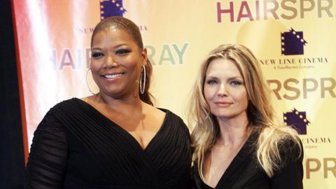 Queen Latifah and Michelle Pfeiffer pose on a carpet in black outfits in front of a banner that says Hairspray.