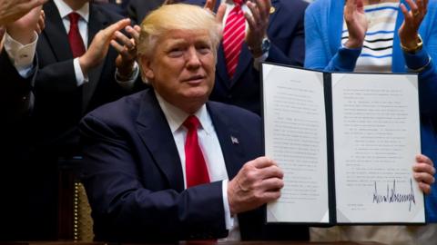 Donald Trump holds up an executive order while people behind him clap