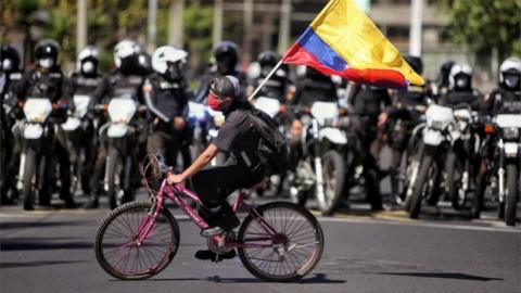 A protester riding a bike waves a flag in front of a row of policemen in Quito