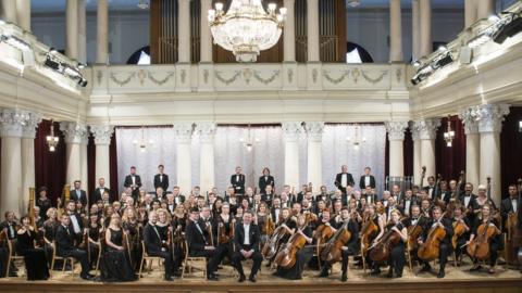 The National Symphony Orchestra of Ukraine posing with their instruments on stage in 2018. They are dressed in formal black tie and the building is ornate with a large glass chandelier above them.