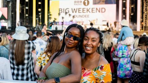 Fans during 鶹Լ Radio 1's Big Weekend at Camperdown Park in Dundee. The fans, two young black women, are pictured at a barrier in front of the stage, a crowd gathering behind them. The woman on the left wears large sunglasses and a strapless green top while her friend wears a bright orange printed blouse. They're pictured outside in the sun, there is no act on stage just a sign saying '鶹Լ Radio 1's Big Weekend Dundee'