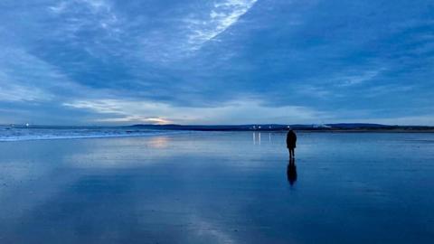 Deep blue sky is reflected in the water as a person walks across a wide beach at dawn