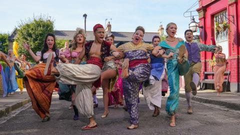 (left to right) Natalie Cassidy as Silly Girl, Maisie Smith as Silly Girl, Zack Morris as Aladdin, Lorraine Stanley as the Genie, Shona McGarty as Belle, Tilly Keeper as Jasmine and Stephen Rahman-Hughes as the Tavern Keeper, as the cast of Eastenders perform a medley of Disney songs in Albert Square for Children In Need.