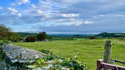 Blue sky with layers of grey, blue and white cloud sit over a field with rolling hills behind and a stone wall in the foreground