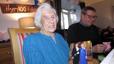 Woman with white hair wearing a blue dress sitting at a table with "Happy 100th Birthday" sign behind her