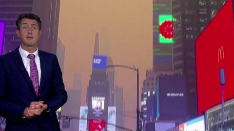 Chris Fawkes standing in front of a shot of Times Square, New York, with the sky coloured brown from the smoke in the air