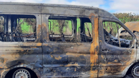 The minibus pictured from the side, completely burnt out, with barely any of its white paint left
