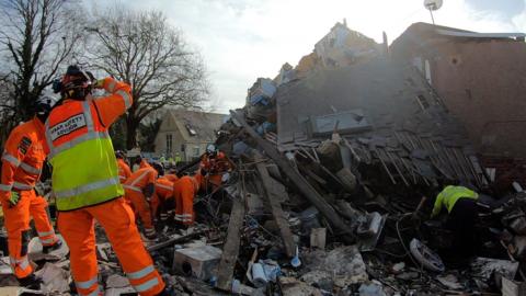 Emergency workers are stood amidst the rubble of the collapsed house. The upper floor of the house is exposed and leaning down to the ground while officers remove rubble.