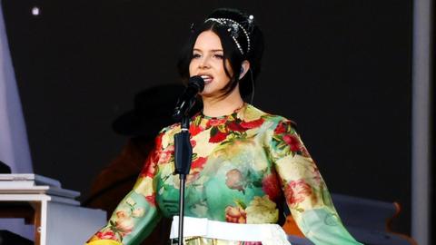 Lana Del Rey singing on stage, she is wearing a green floaty dress and her arms are open. She is wearing her black hair up.