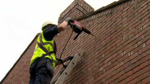 Man in hi-vis drills a hole in the outside of a house