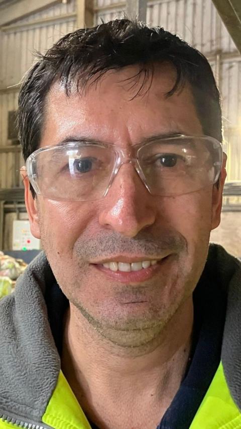 Antonio Rodrigues smiles at the camera. He has short black hair and wears a high vis coat and protective clear glasses.