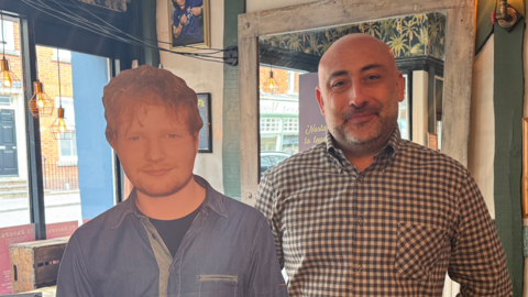 Damon Jeffery wearing a cheque shirt while standing next to an Ed Sheeran cardboard cut out in his pub. 