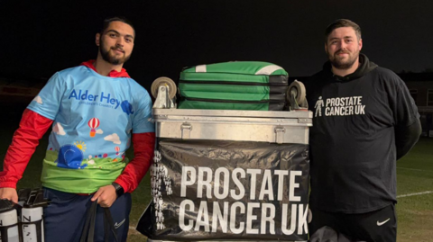 Two men next to a silver case full of football equipment. There is a banner reading Prostate Cancer UK. the man on the left has a sky blue shirt with Alder Hey written on. He has a red undershirt. The man on the right is wearing a black hoodie with Prostate Cancer UK on it in white lettering