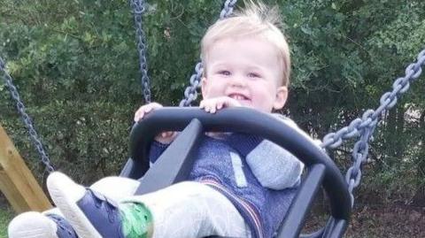 Louis Rogers, aged about one year old, smiles to the camera as he plays on a swing