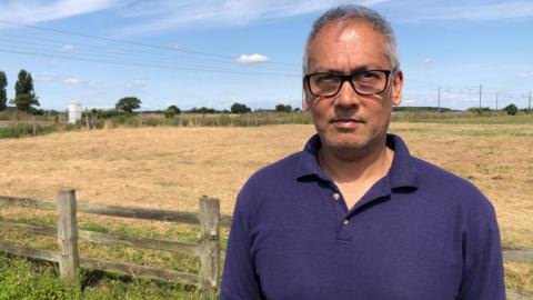 A middle aged man in a blue polo shirt stands in-front of a fence and field of crops