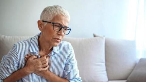 A woman with short grey hair is sitting on the sofa and is pressing both her hands to her chest. Her facial expression suggests she has pain in her chest. She has glasses on and is wearing white shirt with blue stripes. 