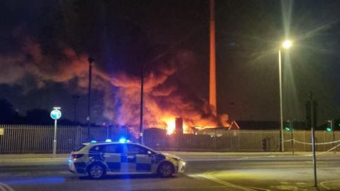Scene of the fire. A police car with blue lights flashing, with smoke and flames seen in the background.