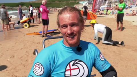 Cam Treble wears a blue Surfing England top over a wetsuit and smiles into the camera 