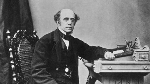 A Victorian black and white photograph of Thomas Cook. He's wearing a smart black tailored coat, and has his left arm on a ornate carved wooden desk.