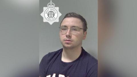 A police mugshot of a young man with short dark hair and glasses, wearing a dark blue t-shirt. The West Mercia Police badge has been superimposed in the top left corner.