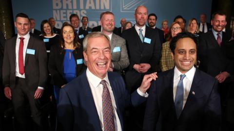 Nigel Farage stands with his hand on the shoulder of Zia Yusuf. Behind them are  the councillors who have defected to Reform UK 