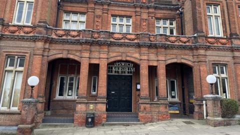 The exterior of Ipswich Museum. It is a large red building with the name of the museum above its entrance.