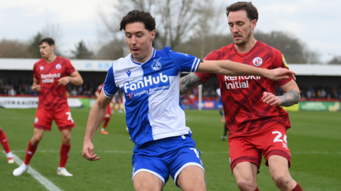 A photo showing two football players competing for the ball. One is wearing a blue and white shirt with blue shorts, the other is wearing an all red kit 