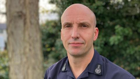 Rick Hylton wearing a navy shirt in front of a tree and a bush. He has a shaved head and looks serious.