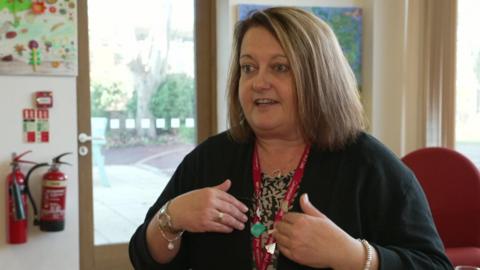 A woman with short, brown hair, wearing a black cardigan, and a red hospice lanyard around her neck, looks to the left of the camera, with her hands to her chest as she is talking. 