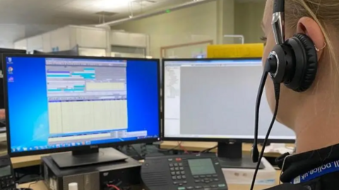 A woman in uniform with a headset looks at two computer screens.