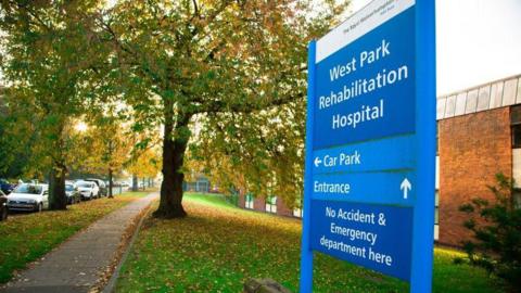 General view of a blue hospital sign which reads West Park rehabilitation hospital twp large trees, a grass verge, a path and a row of cars are also visible